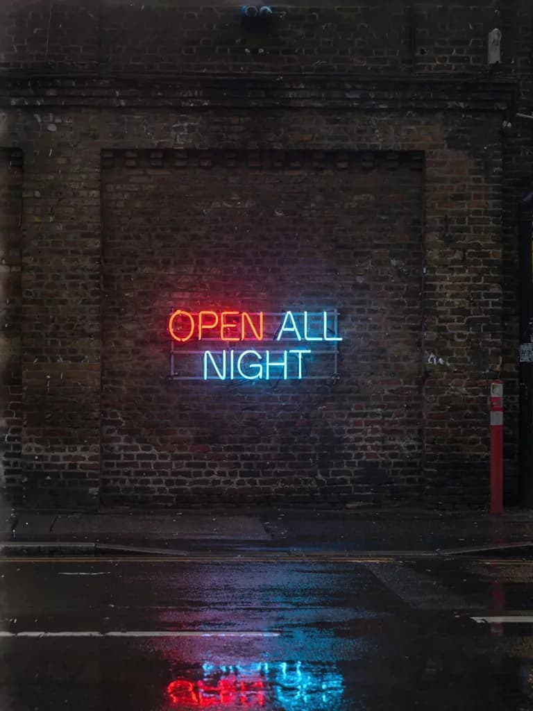 Night street photograph of a glowing neon sign reading "OPEN ALL NIGHT" mounted on a rainy brick wall, reflections on wet pavement